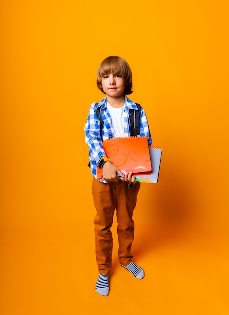 Handsome 7 year old boy with backpack holding books on yellow background for education concept