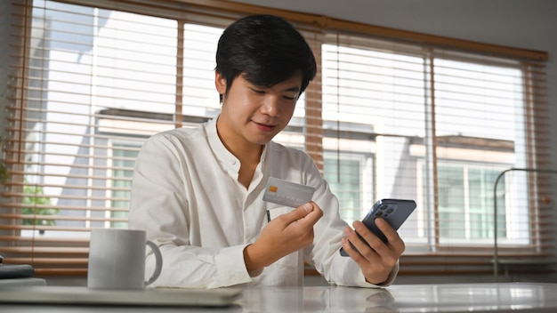 Handsome adult asian man consumer holding credit card and smartphone shopping online or doing online banking transaction