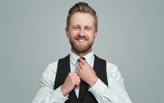 Handsome adult bearded well dressed male in elegant formal clothes adjusting necktie and looking at camera with friendly smile against gray background