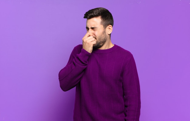 Handsome adult blond man feeling disgusted, holding nose to avoid smelling a foul and unpleasant stench