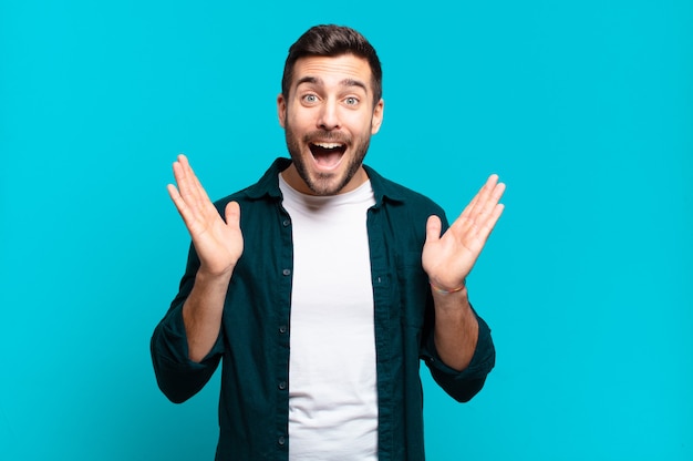 Handsome adult blond man looking happy and excited, shocked with an unexpected surprise with both hands open next to face