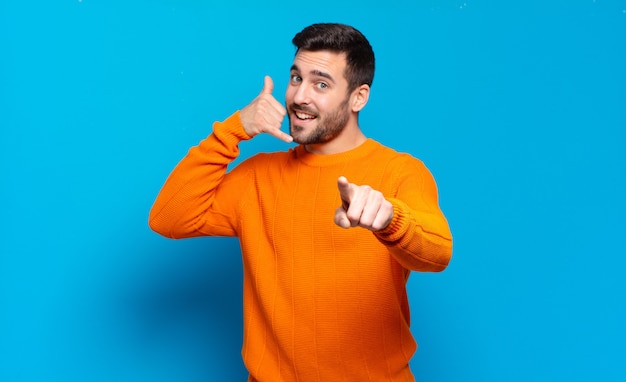 Handsome adult blond man smiling cheerfully and pointing to camera while making a call you later gesture, talking on phone