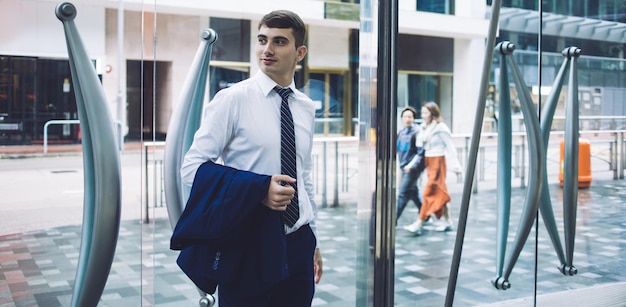 Foto bel maschio adulto in abiti formali con giacca a portata di mano che entra nella luminosa hall dell'edificio per uffici e distoglie lo sguardo