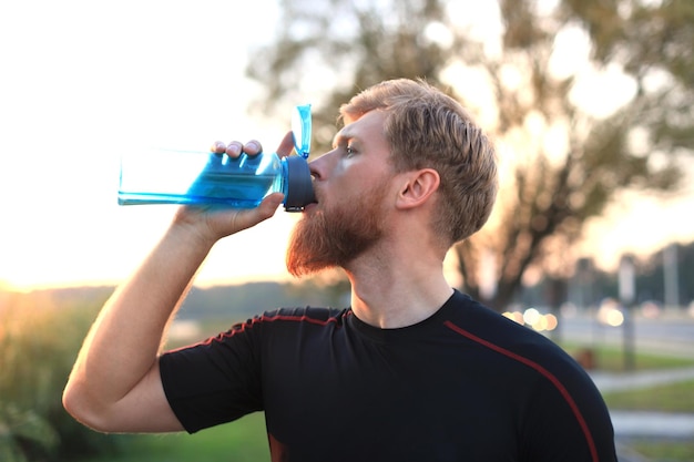 夕焼けや日の出の外に立っている間、フィットネスボトルから水を飲むハンサムな大人の男。ランナー