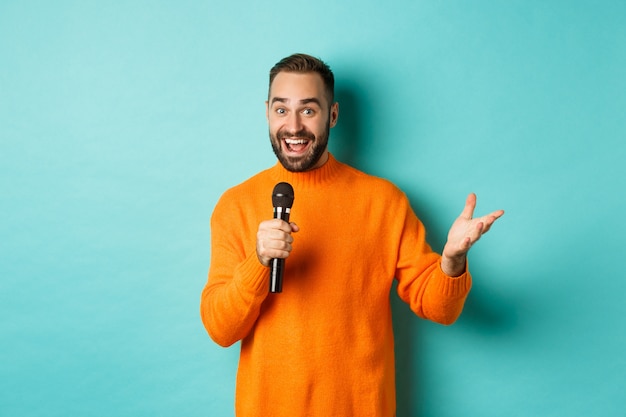 Handsome adult man perform song, singing into microphone, standing against turquoise background.