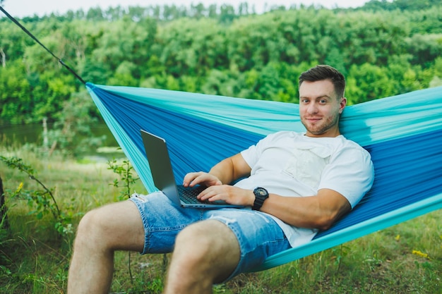 Handsome adult man relaxing in hammock while working on laptop during day hike outdoors Recreation in nature with remote work