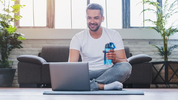 Красивый взрослый мужчина с ноутбуком и питьевой водой во время перерыва во время тренировки дома