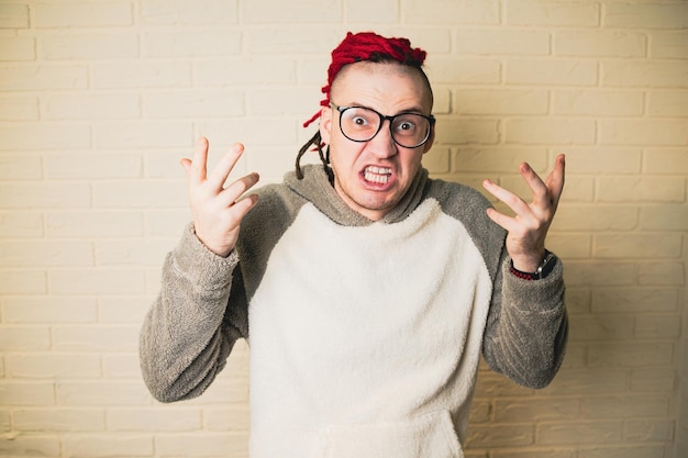 A handsome adult man with glasses and red dreadlocks is angry against a white brick wall