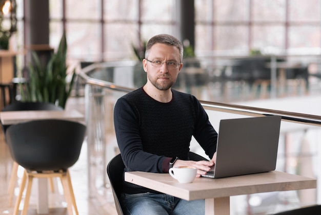 Handsome adult man with glasses works on a laptop in a cafe