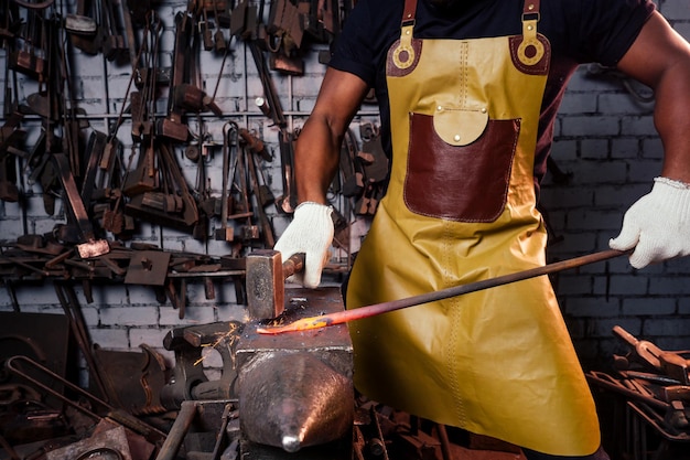 革のエプロンを身に着けているworkshopwearingで働くハンサムなアフリカ系アメリカ人の鍛冶屋の男性労働者