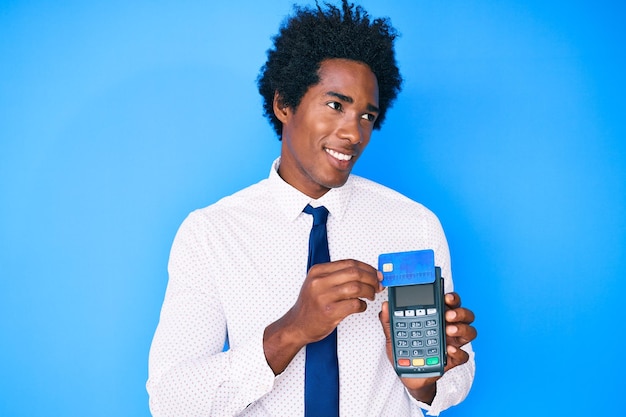 Handsome african american business man with afro hair holding dataphone and credit card smiling looking to the side and staring away thinking