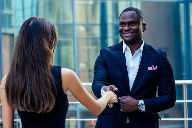 Красивый афроамериканец в черном деловом костюме пожимает руку партнеру-бизнесмену, городскому пейзажу, стеклянным офисам, фону, командной работе и идее успешной сделки
