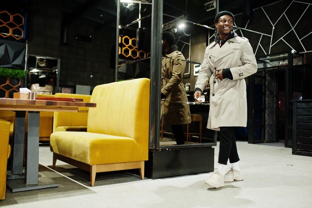 Handsome african american man posing inside night club in black hat and beige coat