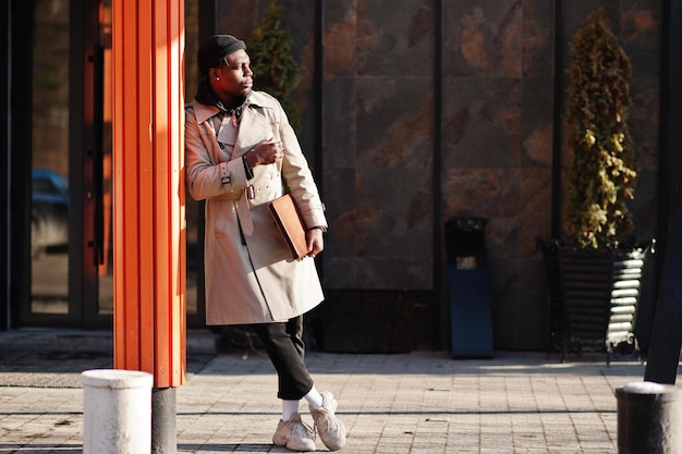 handsome african american man posing outside in black hat and beige coat with folder in hand