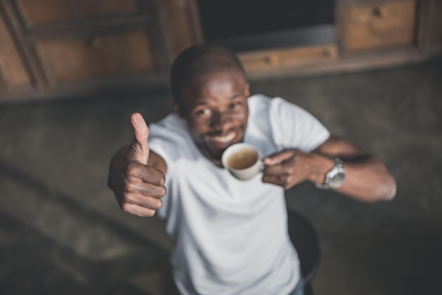 Красивый афроамериканец показывает большой палец вверх и пьет утренний кофе дома