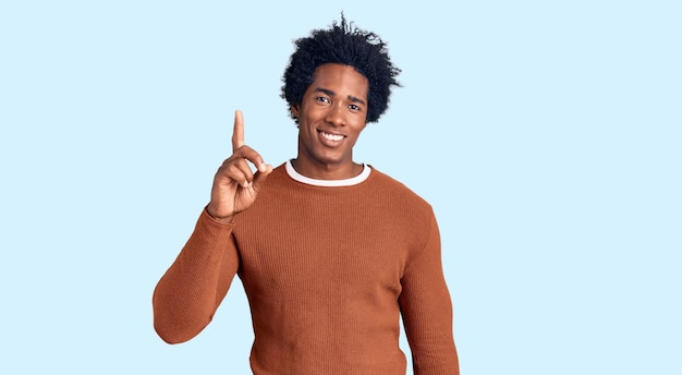 Красивый афроамериканец с афро-волосами в повседневной одежде показывает и указывает пальцем номер один, улыбаясь уверенно и счастливо.