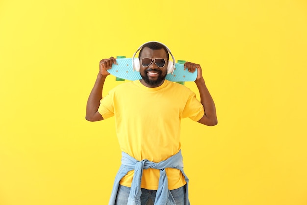 Handsome African-American man with headphones and skateboard on color