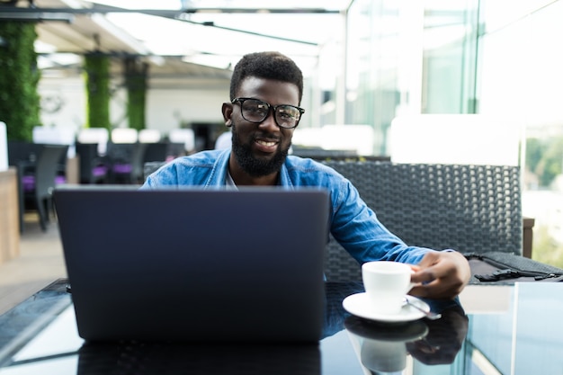 Uomo d'affari africano bello che ha riunione in linea sul computer portatile in un caffè e bere caffè, panorama, spazio della copia