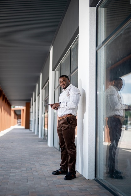 Красивый африканский мужчина держит телефон в руках и улыбается. Сильный мужчина, стоящий у стены бизнес-центра. Бизнесмен в рубашке и джинсах с телефоном у здания.
