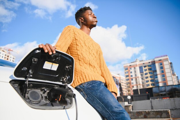 Handsome african man standing near luxury charging electric car and talking on mobile.