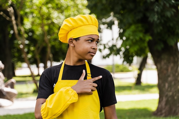 ハンサムなアフリカのティーンエイジャーの料理人が右側を指すシェフの帽子と黄色いエプロンの制服を着た黒い子供料理人が笑顔で右側を指している屋外カフェやレストランのクリエイティブな広告
