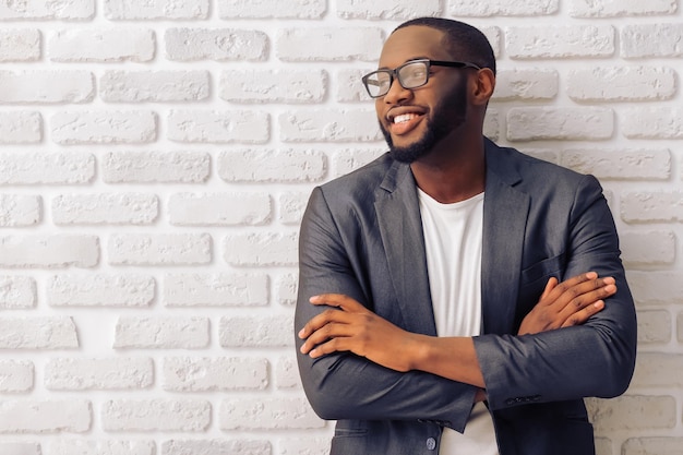 handsome afro american businessman in gray classic jacket and glasses is smiling standing with crossed arms against brick wall