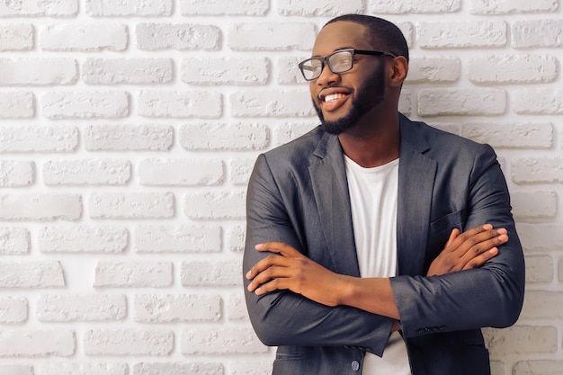 Handsome Afro American businessman in gray classic jacket and glasses is smiling standing with crossed arms against brick wall