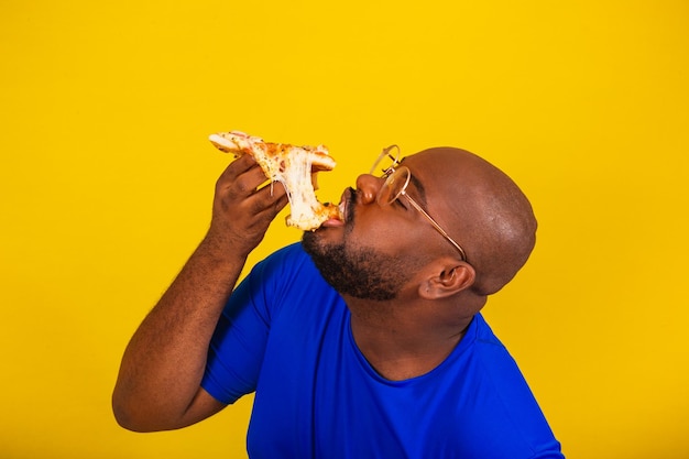 Bell'uomo afro brasiliano con occhiali camicia blu su sfondo giallo mangiare pizza fetta allungamento formaggio formaggio che cade un sacco di formaggio delizioso cremoso da allungare