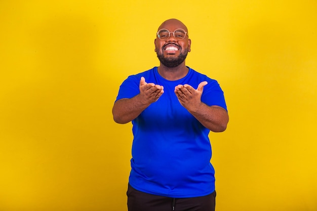 Handsome afro brazilian man wearing glasses blue shirt over yellow background with the hand in the mouth sending kiss kiss greeting friendly see you later goodbye