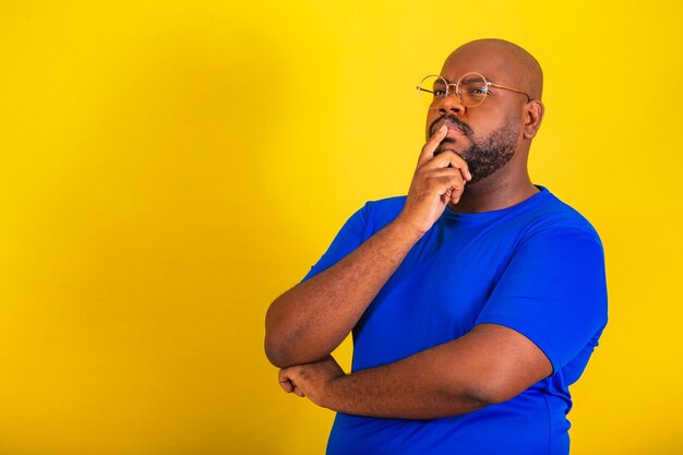 Handsome afro brazilian man wearing glasses blue shirt over yellow background with hands on chin questioning reflection thinking thinking choosing reasoning