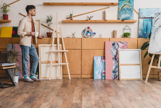 Photo handsome artist standing in spacious light painting studio and holding cup of coffee