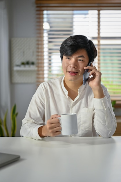 ハンサムなアジア系のビジネスマンが彼のビジネス クライアントと電話で話し、コーヒーをすすりながら