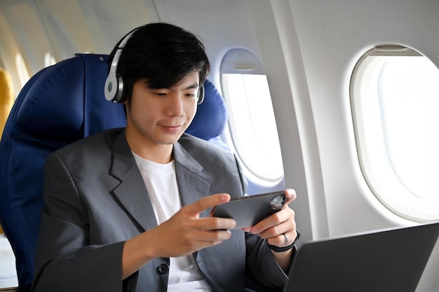 Handsome asian businessman wearing headphone and playing a\
mobile game during the flight
