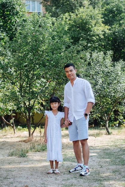 Handsome asian father holding the hand of his daughter standing on a grass in a park, enjoying last warm days of the early fall.