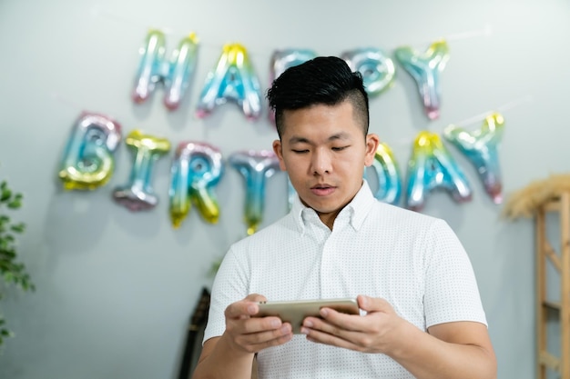 красивый азиатский отец использует свой мобильный телефон, чтобы проверить фотографии на фоне яркого домашнего интерьера с красочным украшением дня рождения.