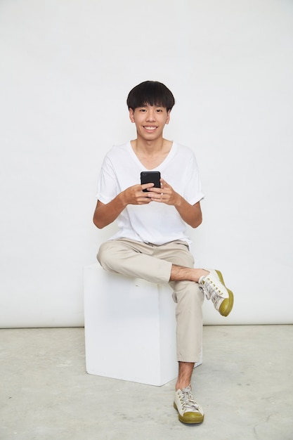 Красивый азиатский парень сидит и пользуется телефоном, изолированным на белой поверхности. копировать пространство
