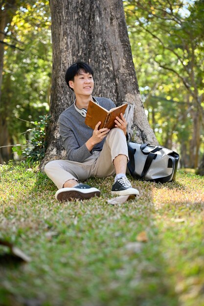 Красивый азиатский студент колледжа читает книгу под деревом в зеленом парке