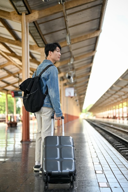 A handsome Asian male traveler with his backpack and suitcase is walking along the platform