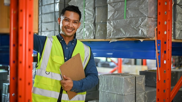 Photo handsome asian male warehouse worker in uniform holding a clipboard paper in warehouse