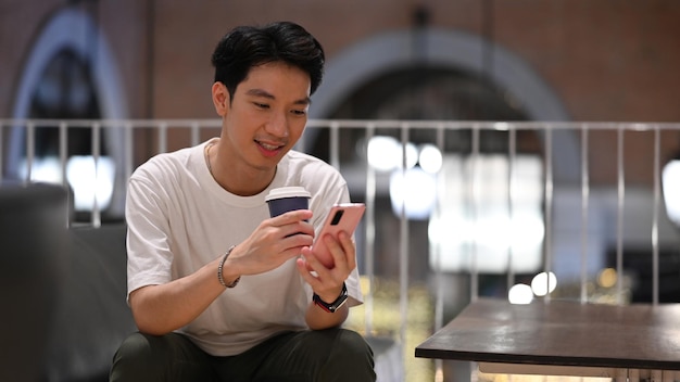 Красивый азиатский мужчина болтает онлайн или проверяет социальные сети на мобильном телефоне в современном кафе