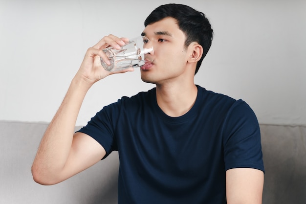 ハンサムなアジア人男性がリビングルームのソファでコップ1杯の水を飲む