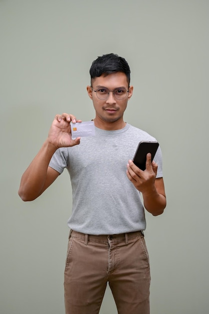 Красивый азиатский мужчина держит свой смартфон и кредитную карту на зеленом студийном фоне