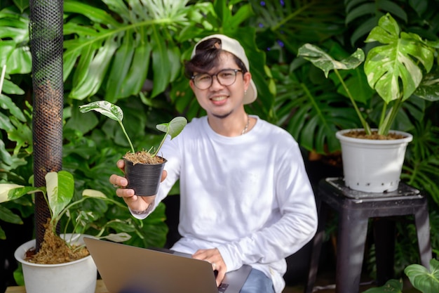 Handsome asian man loves to plant trees sitting in a garden\
monstera is using his laptop computer to market and sell trees\
handsome asian man is interested in doing business selling trees\
online.