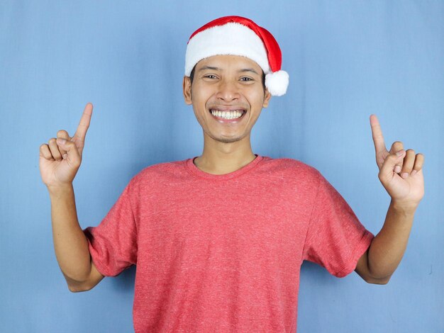 Handsome asian man in red christmas cap pointing fingers isolated over blue background