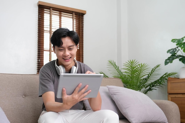 Handsome Asian man using digital tablet while headphones sitting on sofa at home