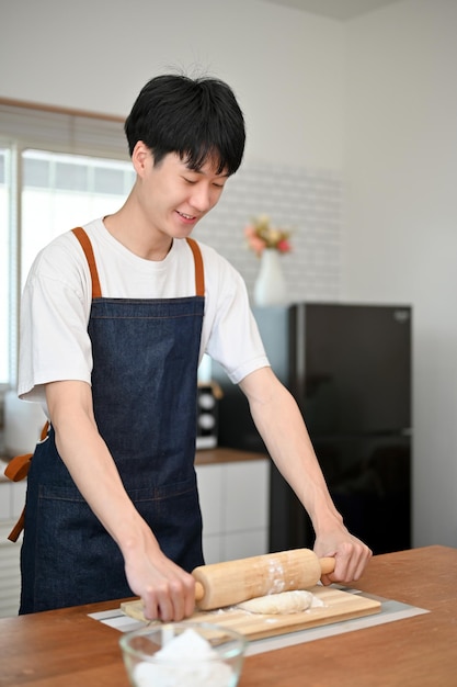 반죽을 만드는 롤링 핀으로 반죽을 반죽하는 앞치마를 입은 잘생긴 아시아 남자