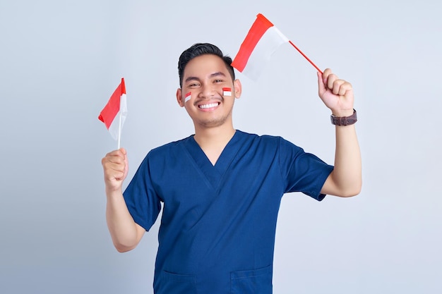 Handsome Asian man wearing blue male nurse uniform holding indonesian flag and celebrating indonesian independence day August 17 isolated on white background studio portrait