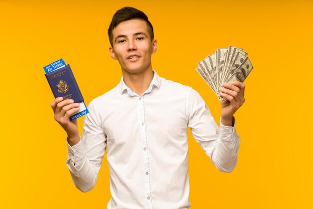 A handsome asian man in a white shirt rejoices in winning the lottery he is holding a passport with air tickets and money dollars on a yellow background image