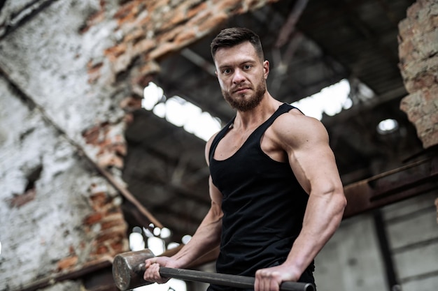 Handsome athlete with hammer in hands. Man on a broken wall background poses to the camera. Abandoned warehouse in the background.