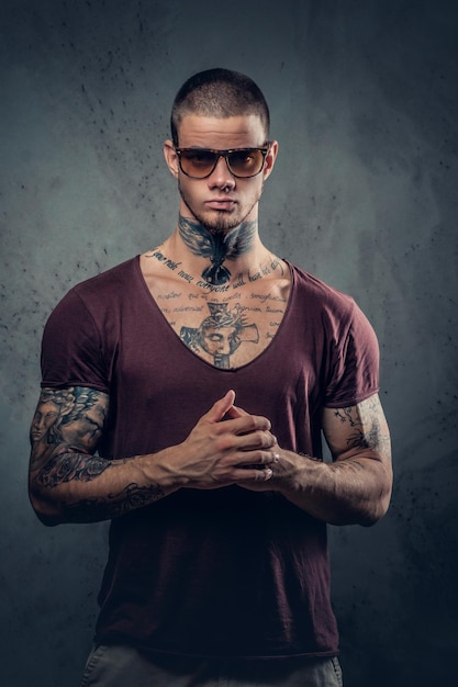 Handsome, athletic male in sunglasses with tattoos on his arms and neck posing over grey artistic background in a studio.